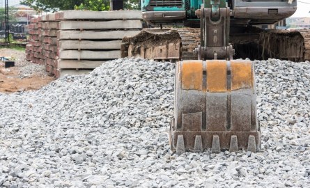 the bucket of a backhoe resting on some gravel with stacks of brick in the background